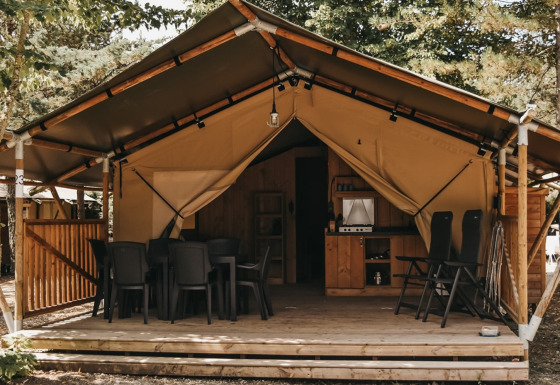 Safari-telt med sanitære faciliteter, udendørs terrasse, spisebord og loungestole under træer.