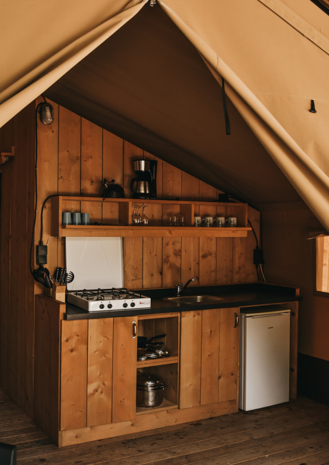 Cocina en tienda safari con paneles de madera, estufa de gas, fregadero y nevera pequeña en Camping Lucherino.