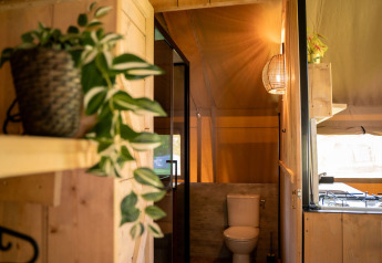 Vista interior de tienda safari con baño, planta verde y decoración de madera en Jocomo Parc, Bélgica.