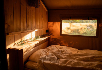 Sunlight illuminates a cozy wooden bedroom with rumpled bedding in a safari tent at Jocomo Parc, Belgium.