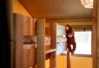 Interior de tienda safari en Jocomo Parc, Bélgica, con literas de madera y un mono de peluche colgando.