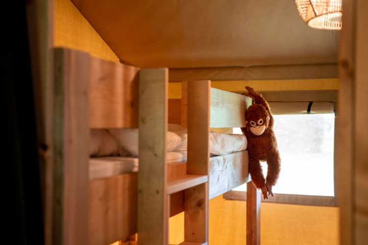 Interior de tienda safari en Jocomo Parc, Bélgica, con literas de madera y un mono de peluche colgando.