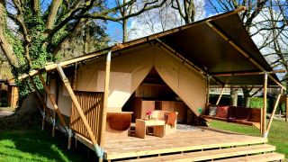 Safari tent met houten veranda en zitgedeelte onder hoge bomen op een zonnige dag in de natuur.