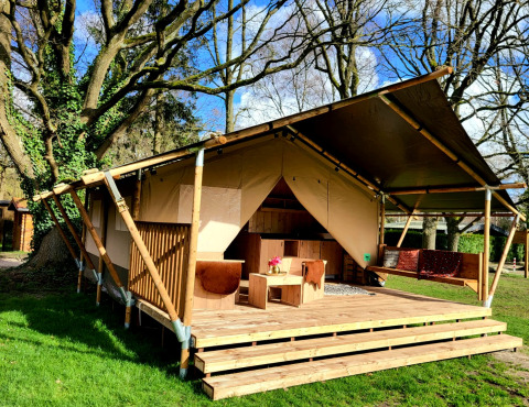 Safari tent with wooden deck and seating area, set under tall trees on a sunny day in nature.