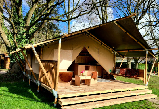 Safari tent met houten veranda en zitgedeelte onder hoge bomen op een zonnige dag in de natuur.