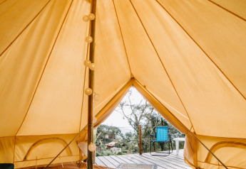 Vista interior de una tienda safari Belltent con cama, luces decorativas y terraza exterior con silla.