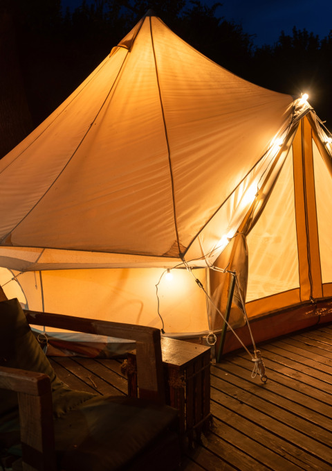 Tienda safari iluminada llamada Belltent en Camping Lucherino, Italia, sobre tarima de madera por la noche.