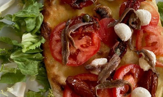 Pizza au pain plat italien garnie de tomates, mozzarella, anchois et salade, servie sur une assiette blanche.