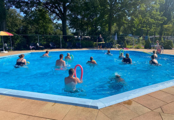 Folk deltager i vandgymnastik i en udendørs pool på en glampingplads omgivet af træer om sommeren.