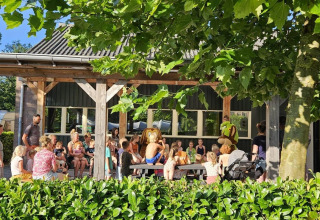 A group of children and adults gather outside under a tree at a glamping site on a sunny day.