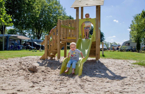 Børn leger på en rutsjebane på en legeplads i sand, omgivet af campingvogne i Den Blanken feriepark.