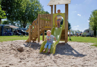 Børn leger på en rutsjebane på en legeplads i sand, omgivet af campingvogne i Den Blanken feriepark.