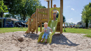 Niños jugando en un tobogán sobre arena, rodeados de caravanas en el parque vacacional Den Blanken.