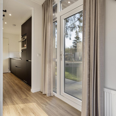 Modern lodge interior at Bospark Ede, Netherlands, with large windows, kitchen and beige curtains.