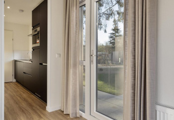 Modern lodge interior at Bospark Ede, Netherlands, with large windows, kitchen and beige curtains.