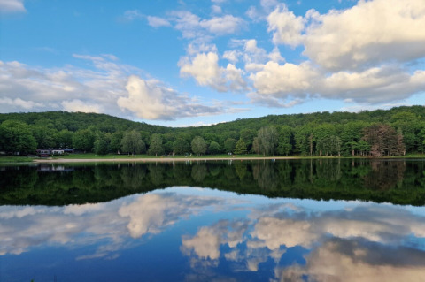 Vista del lago en Camping Montard en Nouvelle-Aquitaine, Francia, con árboles verdes y nubes reflejadas en el agua.
