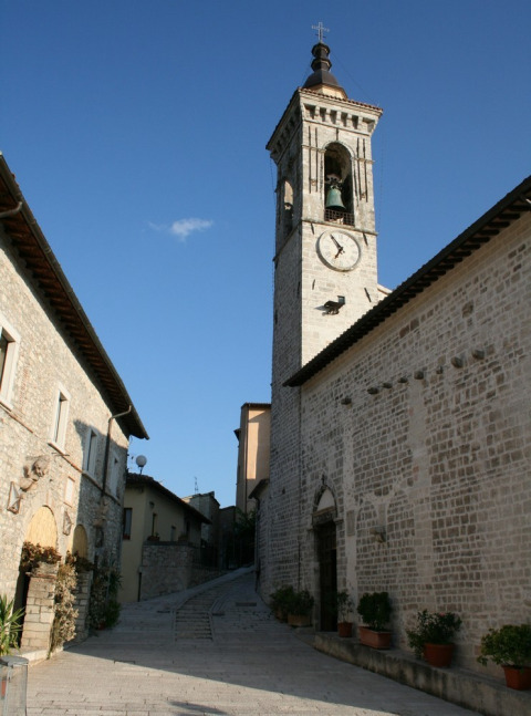 Foto de una torre con campana y casas antiguas de piedra en una calle estrecha de Umbría, Italia, con cielo azul.