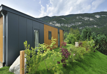 Lodge de montagne moderne en bois et métal sur pelouse en pente, entouré de végétation et de montagnes.