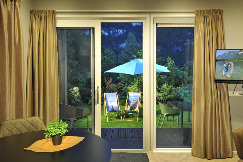 Zicht vanuit de woonkamer van Mountain Lodges op de tuin met ligstoelen en parasol door grote glasdeuren in de avond.