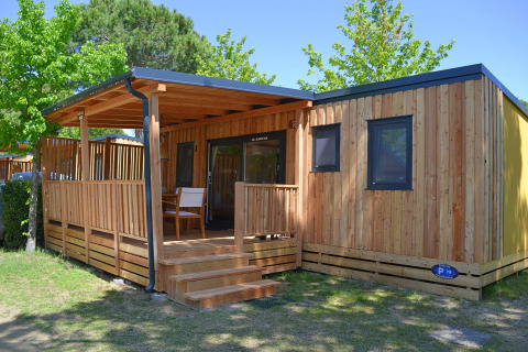 Cabaña SunLodge Sabai de madera con porche cubierto, escalones y sillas en un entorno verde y arbolado.