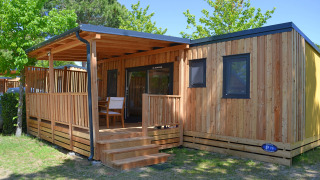 Cabaña SunLodge Sabai de madera con porche cubierto, escalones y sillas en un entorno verde y arbolado.