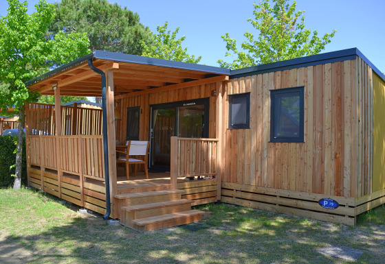 Cabaña SunLodge Sabai de madera con porche cubierto, escalones y sillas en un entorno verde y arbolado.