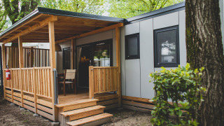 Entrada a SunLodge Sabai, con una moderna cabaña, porche de madera cubierto y sillas, rodeada de árboles.