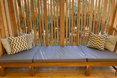 Banc en bois extérieur avec coussins gris et oreillers à motifs à SunLodge Babul, entouré de lattes.