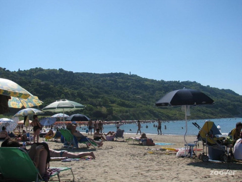 Beach scene at Camping Podere sei Poorte in Marche, Italy, with visitors relaxing under umbrellas and green hills.