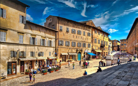 Plaza animada en el Camping Mar y Sierra, parque vacacional en Marche, Italia, con edificios históricos y gente.