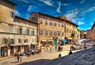 Place animée au Camping Mar y Sierra dans les Marches, Italie, avec bâtiments anciens et nombreux visiteurs.