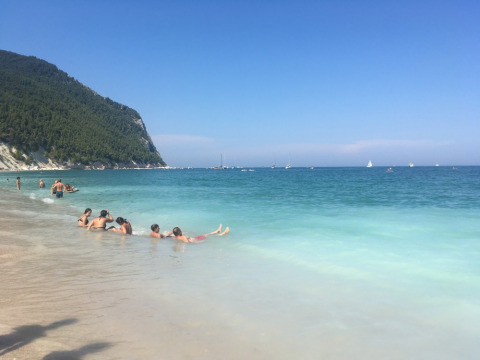Playa en Camping Mar y Sierra, Marche, Italia, con bañistas, mar turquesa, colina y barcos lejanos.