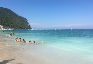 Beach scene at Camping Mar y Sierra in Marche, Italy, with swimmers, turquoise water and boats offshore.