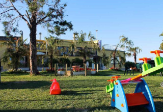 Playground with colorful equipment and lawn in front of holiday park buildings at Camping Mar y Sierra, Italy.