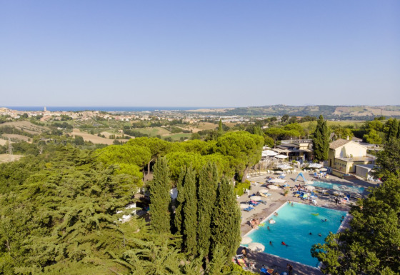 View of Camping Mar y Sierra in Marche, Italy, featuring a large pool, lush trees, and rolling hills landscape.