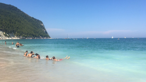 People relaxing on the sandy beach near Stacciola, Marche, Italy, with turquoise water and green hills.