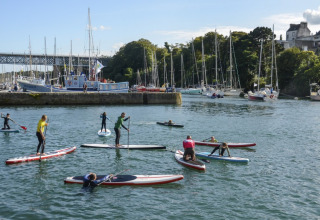 Folk nyder stand up paddleboarding på vandet nær lystbåde og både ved Camping de Kerleyou i Bretagne.