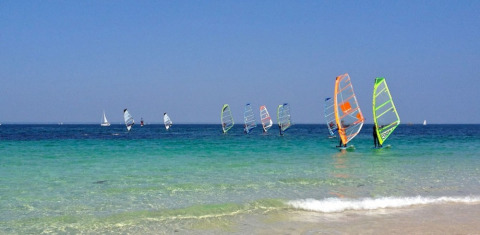 Windsurfistas y veleros en el agua cristalina junto a Camping de Kerleyou, un parque vacacional en Bretaña.