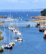 View of Douarnenez harbor in Brittany, France, showing sailboats, lush greenery, and the open sea.