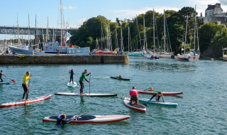 People paddleboarding in the harbor of Douarnenez, Brittany, with sailing boats and buildings in the background.