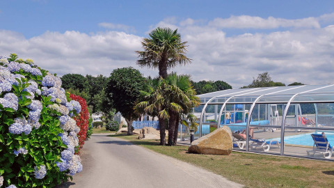 Camino en Camping de Kerleyou, Bretaña, Francia, con flores, palmeras y una piscina cubierta al aire libre.