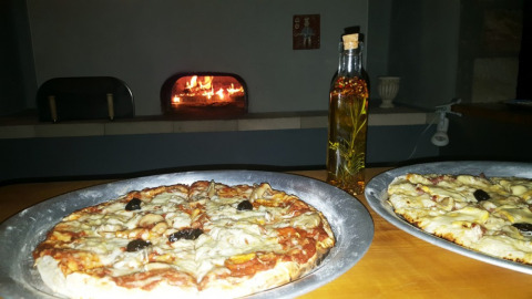 Dos pizzas en bandejas plateadas con una botella de aceite y un horno de leña al fondo en Camping Saint Amand.