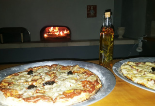 Dos pizzas en bandejas plateadas con una botella de aceite y un horno de leña al fondo en Camping Saint Amand.