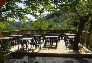Udendørs terrasse med borde og stole under træer på Camping Saint Amand i Auvergne-Rhône-Alpes, Frankrig.