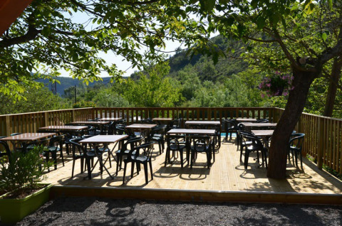 Terraza exterior con mesas y sillas bajo árboles en Camping Saint Amand, Auvergne-Rhône-Alpes, Francia.