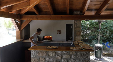 Hombre horneando pizzas en horno de piedra al aire libre bajo techo de madera en Camping Saint Amand, Auvergne-Rhône-Alpes, Francia.