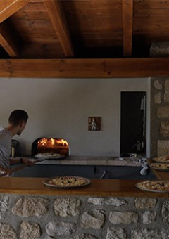 Uomo che cuoce pizze in un forno a pietra all'aperto sotto un tetto di legno al Camping Saint Amand, Auvergne-Rhône-Alpes, Francia.