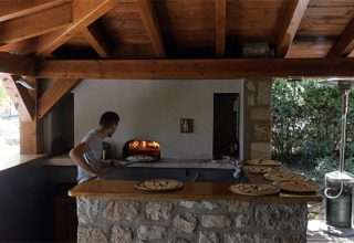 Mann backt Pizza in einem Außen-Steinofen unter einem Holzdach auf Camping Saint Amand, Auvergne-Rhône-Alpes, Frankreich.