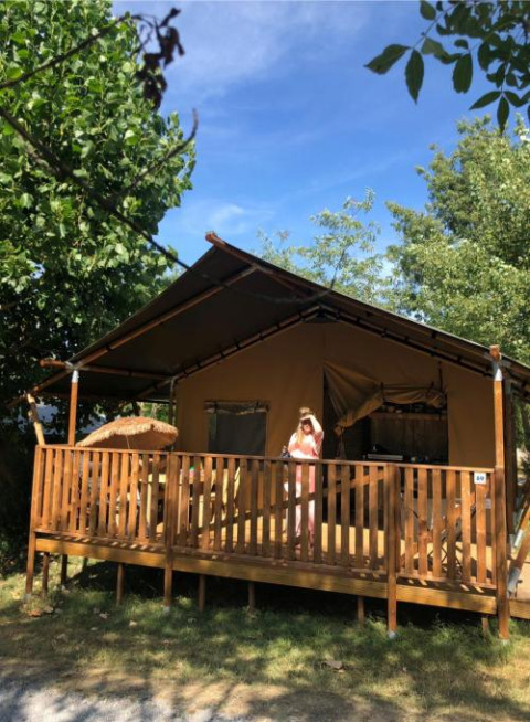 Tienda safari con instalaciones sanitarias en Camping Saint Amand, Francia, persona de pie en el porche soleado.