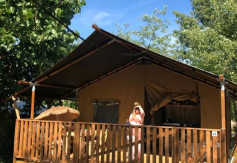 Tienda safari con instalaciones sanitarias en Camping Saint Amand, Francia, persona de pie en el porche soleado.